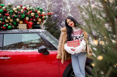 Güzel genç kız kışlık giysiler giyiyor kırmızı arabanın yanında hediye kutusu tutuyor çatıda süslü Noel ağacı, tatil ve yeni yıl konsepti var..