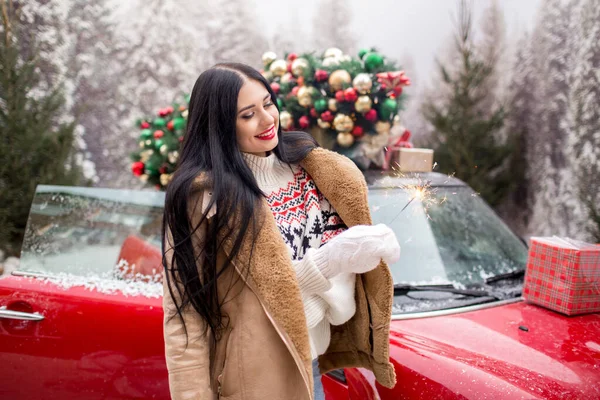 Piękna młoda dziewczyna nosi zimowe ubrania gospodarstwa ognie w pobliżu czerwonego samochodu z ozdobione choinki na dachu, wakacje i koncepcja nowego roku. — Zdjęcie stockowe