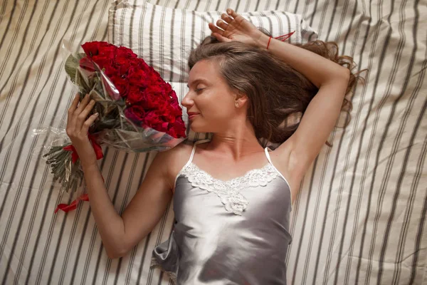 Selamat pagi wanita muda yang menarik di tempat tidur memegang karangan bunga dari seratus mawar merah . — Stok Foto