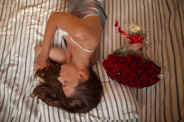 Selamat pagi wanita muda yang menarik dengan karangan bunga dari ratusan mawar merah sedang tidur di rumah . — Stok Foto