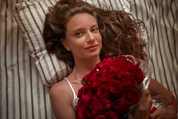 Good morning Attractive young woman in bed is holding bouquet of hundred red roses. — Stockfoto