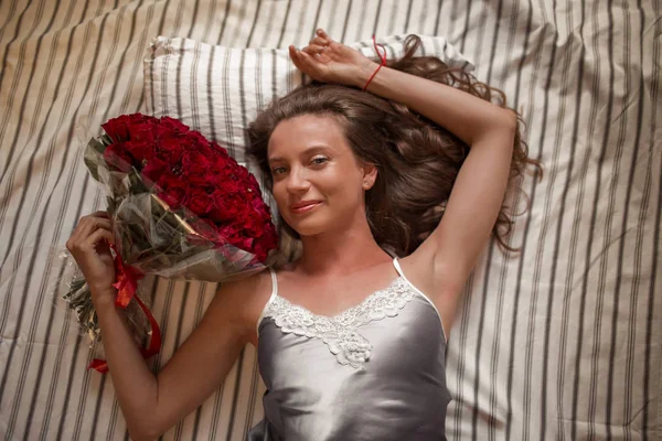 Good morning Attractive young woman in bed is holding bouquet of hundred red roses. — Stockfoto