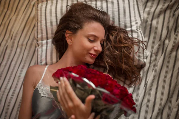 Buongiorno Giovane donna attraente a letto sta tenendo bouquet di cento rose rosse . — Foto Stock