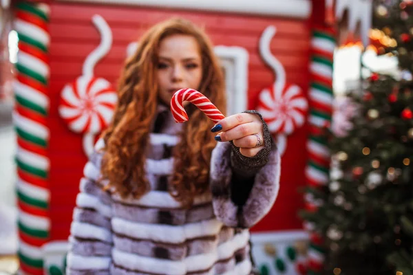 Outdoor portrait of young beautiful redhead happy smiling girl with traditional xmas lollipop. Festive Christmas house on background. — Stock Photo, Image
