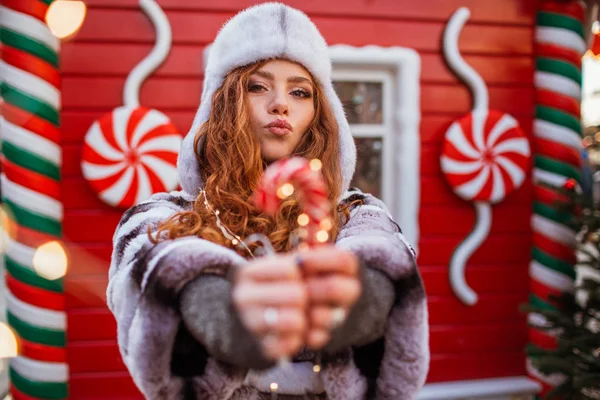 Outdoor portrait of young beautiful redhead happy smiling girl is wearing hat with traditional xmas lollipop. Festive red Christmas house on background. — Stock Photo, Image