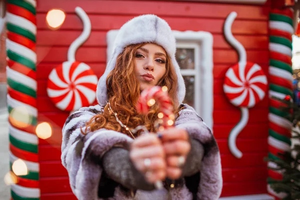 행복하게 웃는 젊은 빨간 머리 소녀가 전통적 인 Xmas Lollipop 모자를 쓰고 야외에서 있다. 배경에는 축제같은 붉은 크리스마스 하우스. — 스톡 사진