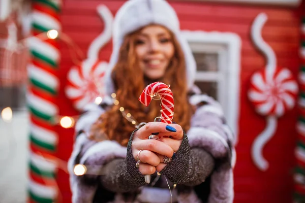 Outdoor portrait of young beautiful redhead happy smiling girl is wearing hat with traditional xmas lollipop. Festive red Christmas house on background. — Stock Photo, Image