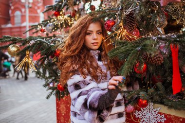 Açık havada güzel kızıl saçlı mutlu bir kızın portresi süslenmiş bir Noel ağacının yanında kürk giyiyor..