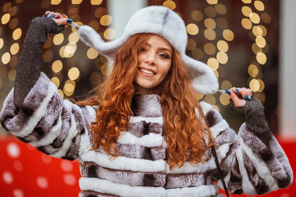 Outdoor portret van jonge mooie roodharige vrolijke lachende meisje draagt bont warme hoed. Feestelijke kerstverlichting op achtergrond. — Stockfoto