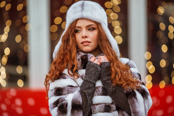 Retrato ao ar livre de jovem linda ruiva feliz sorrindo menina está vestindo chapéu de pele quente. Luzes festivas de Natal no fundo . — Fotografia de Stock