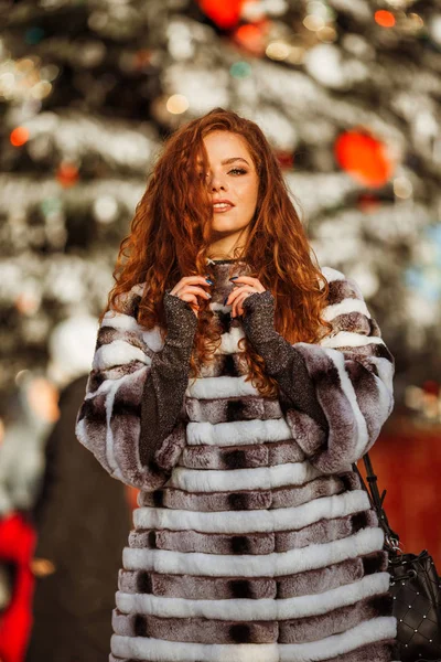 Outdoor portrait of young beautiful redhead happy smiling girl is wearing fur warm hat. Festive Christmas lights on background. — Stock Photo, Image