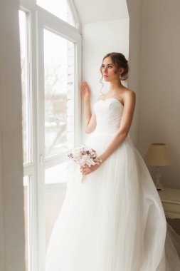 Parlak beyaz stüdyonun penceresinin yanında elinde bir buket çiçek tutan güzel elbiseli genç gelin. Düğün konsepti.