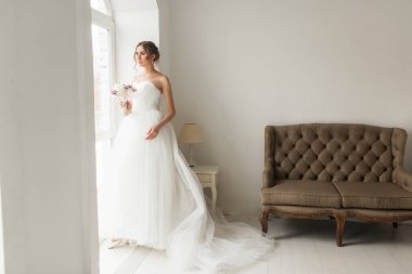 Parlak beyaz stüdyonun penceresinin yanında elinde bir buket çiçek tutan güzel elbiseli genç gelin. Düğün konsepti.