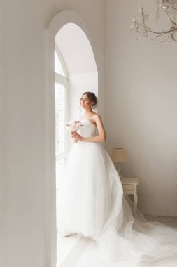 Parlak beyaz stüdyonun penceresinin yanında elinde bir buket çiçek tutan güzel elbiseli genç gelin. Düğün konsepti.