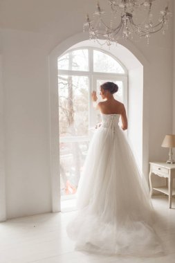 Parlak beyaz stüdyonun penceresinin yanında elinde bir buket çiçek tutan güzel elbiseli genç gelin. Düğün konsepti.