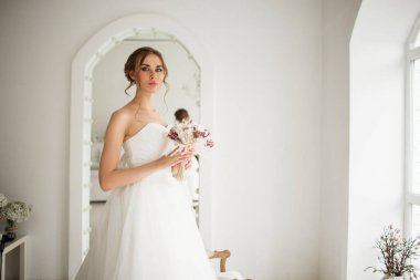 Lüks elbiseli genç gelin parlak beyaz stüdyoda bir buket çiçek tutuyor. Düğün moda konsepti.