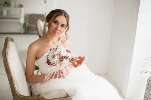 Jonge bruid in een prachtige jurk met een boeket bloemen in helder witte studio. Wedding concept. — Stockfoto