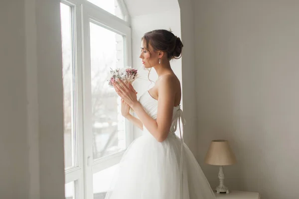 Joven novia en un hermoso vestido sosteniendo un ramo de flores posando cerca de la ventana en un estudio blanco brillante. Concepto de boda . — Foto de Stock