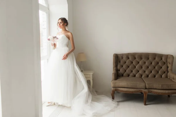 Joven novia en un hermoso vestido sosteniendo un ramo de flores posando cerca de la ventana en un estudio blanco brillante. Concepto de boda . — Foto de Stock