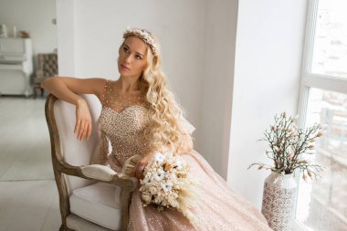 Güzel elbiseli ve kafasında çelenk olan genç gelin parlak beyaz stüdyoda bir buket çiçek tutuyor. Düğün konsepti.