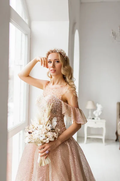 Young bride in a beautiful dress and wreath on her head holding a bouquet of flowers in bright white studio. Wedding concept. — Stock Photo, Image