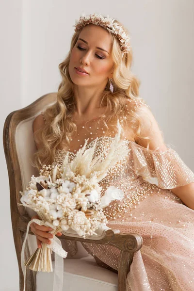 Young bride in a beautiful dress and wreath on her head holding a bouquet of flowers in bright white studio. Wedding concept. — Stock Photo, Image