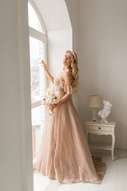 Güzel elbiseli ve kafasında çelenk olan genç gelin parlak beyaz stüdyoda bir buket çiçek tutuyor. Düğün konsepti.