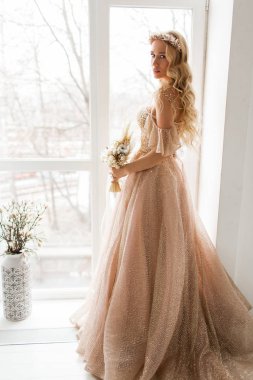 Güzel elbiseli ve kafasında çelenk olan genç gelin parlak beyaz stüdyoda bir buket çiçek tutuyor. Düğün konsepti.