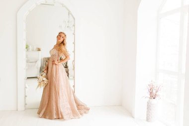 Güzel elbiseli ve kafasında çelenk olan genç gelin parlak beyaz stüdyoda bir buket çiçek tutuyor. Düğün konsepti.