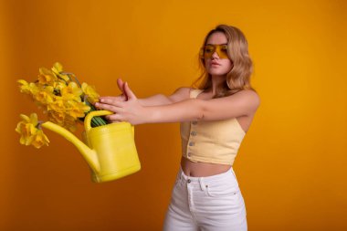 Stüdyodaki güzel kız sarı arka planda sulandırıcı kutusuyla narsist çiçeklerle dolu. Yaratıcı ve moda kavramı.
