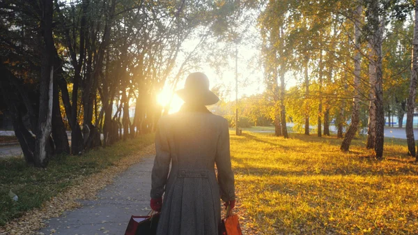 Mature elegant woman in a coat and  hat with their purchases in the park going through the sun at sunset.