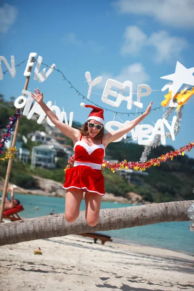 Cute joyful woman jumps in red dress, sunglasses and santa hat on exotic tropical beach. Holiday concept for New Years Cards. Koh Samui, Thailand