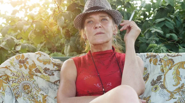 Mature woman in hat sitting in the garden smoking cigarette.