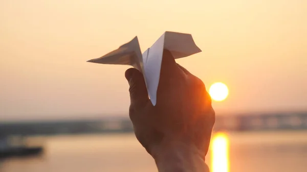 Mans hand of launch paper plane against the sea during sunset with sun flare and reflections in the water in slowmotion, as in childhood.