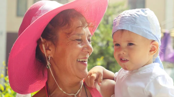 Fashionable happy smiles grandma wearing pink hat holds grandson in sunny summer day at the park