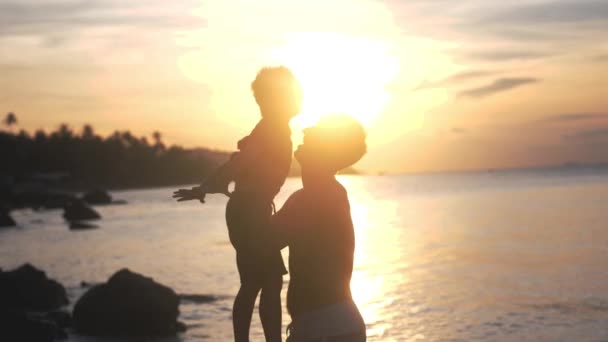 Táta pořád na ruce a hází syna v brýlích na úžasný západ slunce na tropické pláži. Zpomalený pohyb. 3840 x 2160
