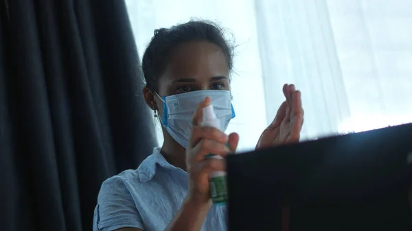 Woman in mask by computer quarantined at home during a pandemic. A woman treats hands with an antiseptic and works on notebook.