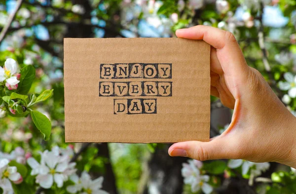 Woman hand holding cardboard card with words Enjoy Every Day