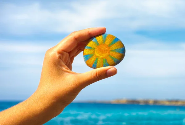 Person hand hold pebble with painted bright sun against blue sky