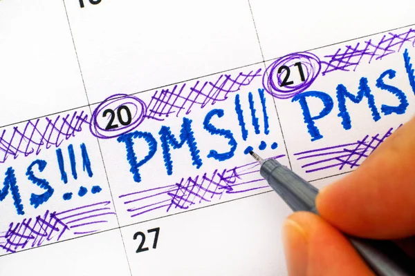 Woman fingers with pen writing reminder PMS in calendar.