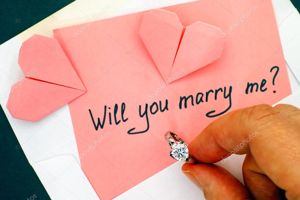 Person hand holding engagement ring. Letter with text Will you m