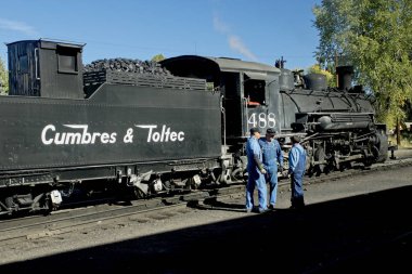 Chama NM, USA, Sept. 19, 2017: Railroad workers chat as a vintage steam locomoitve from the Cumbres & Toltec warms up in the railyard before an autumn excursion. clipart