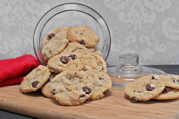 Chocolate chip cookies spilling out of a glass cookie jar.