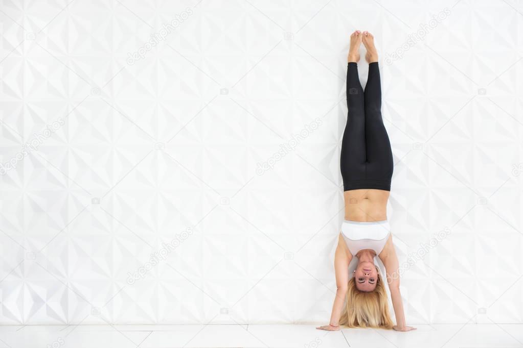 Young blonde woman doing a handstand over a white wall