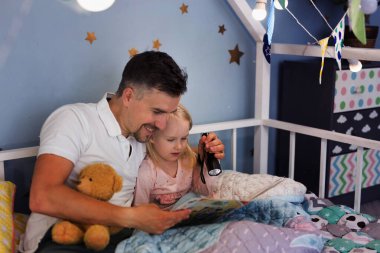 Genç, yakışıklı baba ve kızı yatakta yorganın altında otururken el feneriyle kitap okuyorlar.