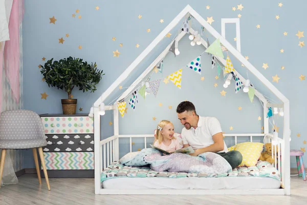 Happy father laughing with his 3 years old girl while sitting on white house bed in children bedroom — Stock Photo, Image