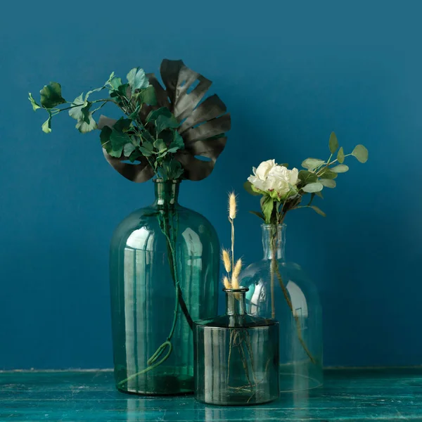 Various artificial flowers and dry plants in transparent vases on the floor against blue wall — Stock Photo, Image