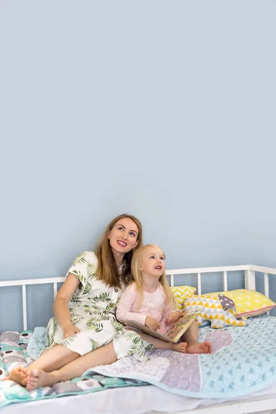 Glückliche junge Mutter und ihre entzückende Tochter schauen auf, während sie zusammen auf dem Bett sitzen — Stockfoto