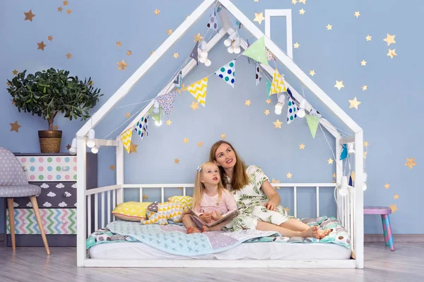 Happy young mother and her cute daughter looking up while sitting in cute house bed — Stock Photo, Image