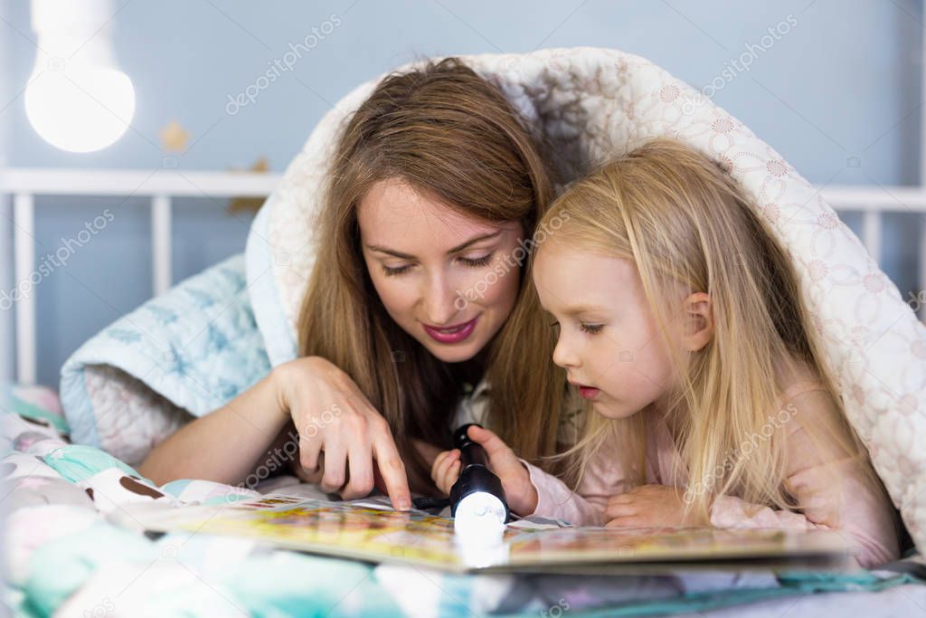 Young mother reading a book using flashlight with her pretty girl while lying down on bed